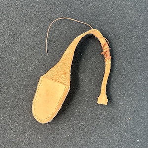 May include: A light brown leather pouch with a curved strap, possibly for a small tool or accessory. The strap is wrapped with copper wire near the end. The pouch is on a dark gray surface.