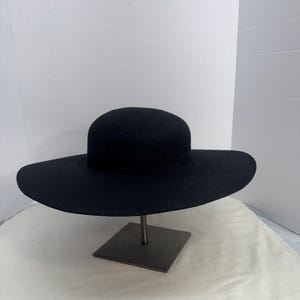 Peut inclure: Un chapeau noir avec un large bord et une couronne arrondie. Le chapeau est présenté sur un support métallique avec une base carrée. Le bord du chapeau est large et la couronne est en forme de dôme arrondi. Le chapeau est fait d'une matière douce.