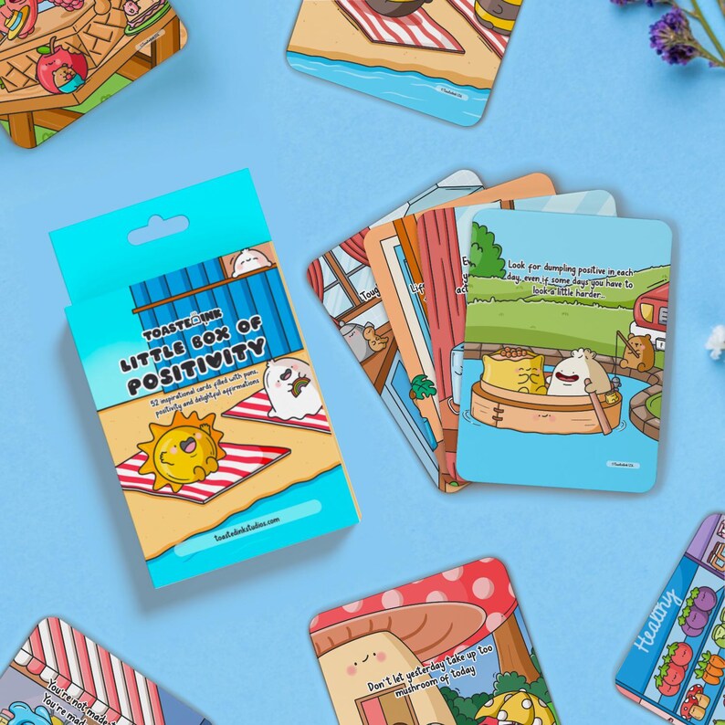 May include: A blue box with the words "TOASTEDINK LITTLE BOX OF POSITIVITY" on the front. The box contains 52 inspirational cards filled with positive and insightful affirmations. The cards feature cute illustrations of cartoon characters.