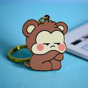 May include: A gold-coloured keychain featuring a cartoon monkey with a brown body, white face, and pink cheeks. The monkey has a grumpy expression and is holding its arms crossed.