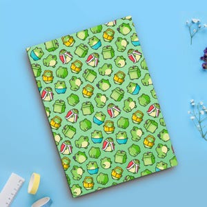 以下が含まれることがあります： 王冠をかぶったカエルが繰り返し描かれた緑色のノート。カエルは緑、黄、赤、青です。背景は薄い緑色です。