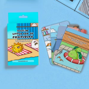 May include: A box containing 52 inspirational cards filled with puns, positivity, and delightful affirmations. The box is blue with a cartoon sun and a ghost on the front. The cards feature illustrations of a turtle, a cat, and a dog.