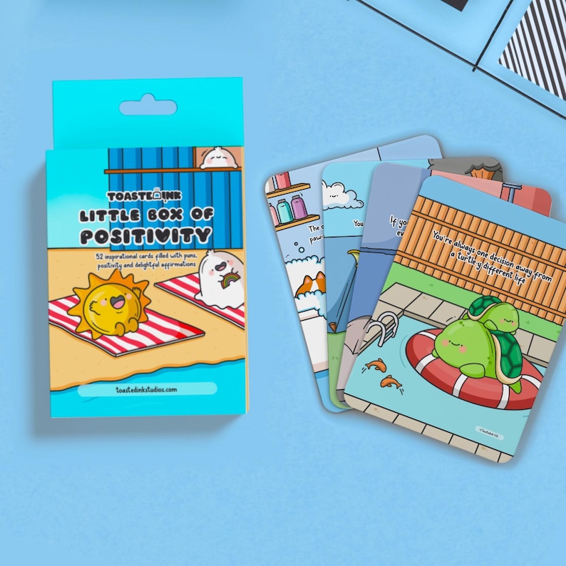 May include: A box containing 52 inspirational cards filled with puns, positivity, and delightful affirmations. The box is blue with a cartoon sun and a ghost on the front. The cards feature illustrations of a turtle, a cat, and a dog.