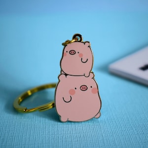 May include: A gold-tone keychain featuring two stacked pink pig charms. The pigs have black eyes, pink cheeks, and black snouts. The keychain has a gold-tone ring.