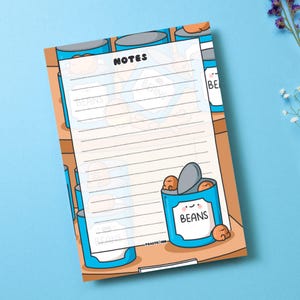 May include: A blue and white lined notepad with a cartoon illustration of a can of beans with a smiling face. The can is labelled "BEANS". The notepad has the word "NOTES" at the top.