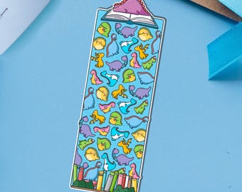 Puede incluir: Marcapáginas rectangular con un patrón de dinosaurios coloridos. El marcapáginas tiene un fondo azul con varios dinosaurios de dibujos animados en tonos amarillos, verdes, rosas y morados. Un pequeño libro abierto con un dinosaurio morado en la parte superior.