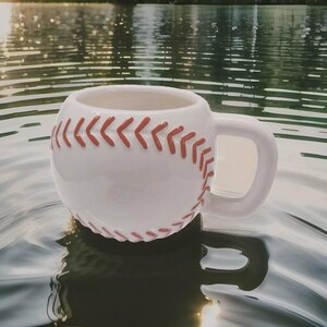 May include: White ceramic baseball mug with red stitching. The mug is floating in a body of water.