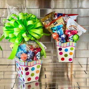 May include: Two gift baskets filled with candy and snacks. The baskets are white with colorful polka dots and striped rims. One basket has a large green bow. The baskets contain lollipops, gummy candies, and chips.
