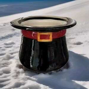 May include: A black ceramic planter shaped like a top hat with a red and gold band around the brim. The planter is sitting on a snowy surface.