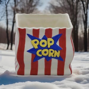May include: A ceramic popcorn container shaped like a classic popcorn bag. The bag is white with red vertical stripes and features the words "POP CORN" in yellow letters on a blue starburst design.
