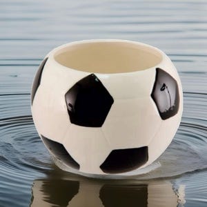 May include: A ceramic soccer ball-shaped container with a white exterior and black pentagonal patches. The interior is a light beige color. The container is sitting in water, creating a reflective surface.