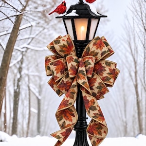 May include: A black metal lamp post with a glowing light, topped with two red cardinal figurines and a dusting of snow. A large, multi-looped decorative bow made of tan fabric featuring a vibrant autumn leaf print is tied around the post.