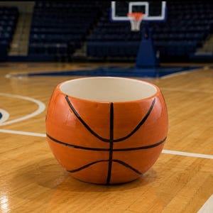 May include: A round ceramic bowl featuring a textured orange surface with black lines, designed to resemble a basketball. The interior of the bowl is smooth and white, providing a contrast to the exterior pattern of the sports-themed container.