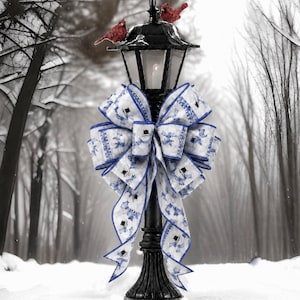 May include: A black metal lamp post topped with two small red cardinal figurines, adorned with a large decorative bow featuring a white and blue snowman pattern. The bow has long, flowing tails and is tied securely around the center of the post.