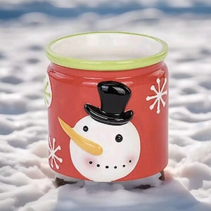 May include: A red ceramic snowman-shaped candle warmer with a green rim and white snowflakes. The snowman has a black top hat, orange nose, and black eyes.