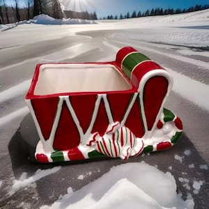 May include: A red and green ceramic Christmas sleigh planter with white trim and candy cane accents. The planter is sitting on a snowy surface.
