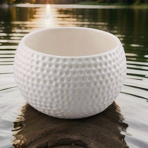 May include: A white ceramic bowl with a textured, dimpled surface, resembling a golf ball. The bowl has a rounded shape and a smooth, off-white interior. The bowl is sitting on a piece of wood, with a blurred background of water and sunlight.