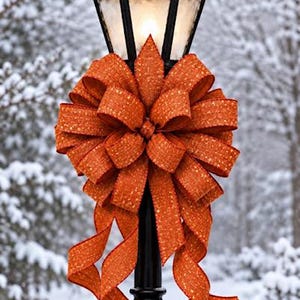 May include: A black metal lamp post topped with two red cardinal bird figurines and a dusting of snow. A large, textured orange ribbon bow with gold flecks is tied around the center of the post, featuring long, flowing tails that drape downward.