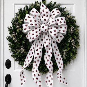May include: A round green boxwood wreath featuring small white berries and a large, multi-looped white ribbon bow. The ribbon is decorated with a repeating pattern of small red ladybugs and hangs down in four long, flowing, curled tails.