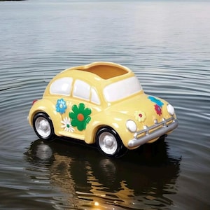 May include: A yellow ceramic Volkswagen Beetle planter with colorful flower decals. The planter has a white interior and is designed to hold plants or flowers.