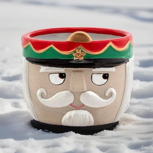 May include: A ceramic nutcracker planter with a red, green, and gold hat. The planter has a white face with a black mustache and beard.