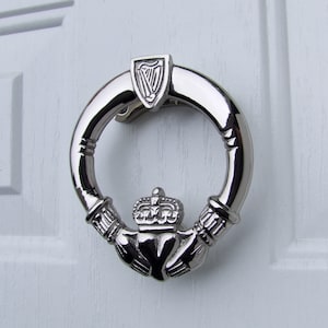 May include: Silver door knocker shaped like a Claddagh ring with a crown, two hands clasped together, and a heart. The ring is attached to a silver plaque with a harp design.