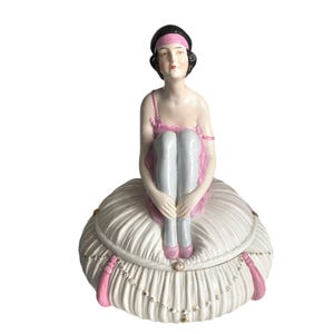 May include: A porcelain figurine of a seated woman in a pink dress and headband, with white stockings and shoes. The figurine sits atop a white, ruffled, lidded box with pink accents and gold-coloured details. A decorative piece.