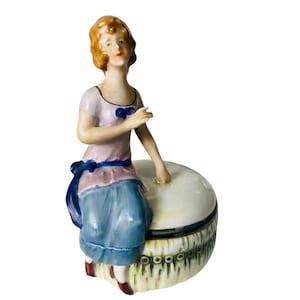Könnte beinhalten: Vintage Porzellanfigur einer sitzenden Frau mit rotbraunem Haar, die ein hellrosa Oberteil mit blauer Schleife und einen blauen Rock trägt. Die Figur sitzt auf einer weißen und cremefarbenen runden Box mit dekorativem Besatz.