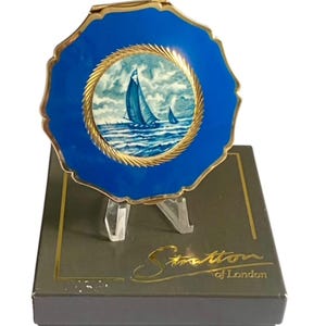 May include: A round Stratton of London compact mirror featuring a vibrant blue enamel border with a scalloped edge and a central circular illustration of sailboats on a choppy sea, framed by a gold-toned rope-textured metal border.