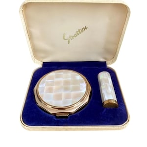 May include: A vintage Stratton vanity set in a cream-colored case with a blue velvet interior. The set includes a round, gold-toned compact with a white mother-of-pearl checkerboard inlay and a matching cylindrical lipstick case with the same inlay.