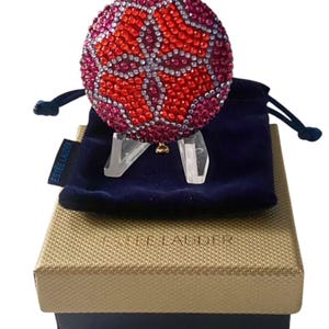 Puede incluir: Un espejo compacto redondo y enjoyado con un diseño floral en tonos rojos, rosas y plateados. El compacto se encuentra sobre un soporte transparente, encima de una bolsa con cordón azul marino y una caja dorada. La caja tiene una base negra y el nombre de la marca.