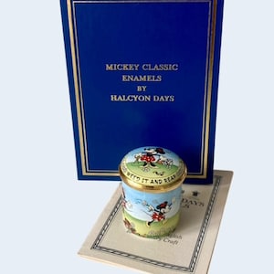 Pode incluir: Uma caixa cilíndrica de esmalte com uma ilustração colorida da Minnie Mouse a fazer jardinagem e o texto "WEED IT AND REAP" na tampa. Inclui uma caixa de apresentação azul com letras douradas "MICKEY CLASSIC ENAMELS BY HALCYON DAYS" e um folheto informativo.