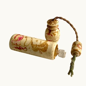Könnte beinhalten: Elfenbeinfarbene antike Parfümflasche mit floralen Designs in Braun und Rot. Die Flasche hat einen durchsichtigen, quadratischen Stopfen und ist mit zwei dekorativen Teilen durch eine braune Kordel und eine grüne Quaste verbunden. Die Teile sind zylindrisch und rund.
