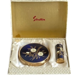 May include: A vintage Stratton powder compact and matching lipstick case set in a presentation box. Both items feature a deep blue enamel finish with a gold-tone floral thistle design. The interior of the box lid displays the red Stratton logo.
