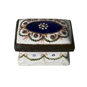 Könnte beinhalten: Eine rechteckige, antike Schmuckschatulle mit Scharnierdeckel. Der Deckel hat ein zentrales blaues ovales Design mit einem weißen Blumenmuster und wird von einem dunklen Metallrahmen umrandet. Die Basis der Box ist weiß mit einem dekorativen, gewellten Rand.