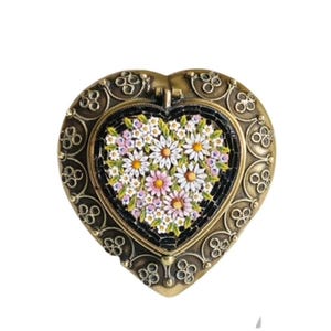 May include: A heart-shaped trinket box featuring an antique brass-toned metal frame with intricate scrollwork. The center inlay displays a mosaic pattern of white and pink daisy-like flowers with yellow centers set against a black background.