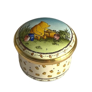 Pode incluir: Uma caixa de joias redonda de porcelana com uma borda de metal dourado. A tampa apresenta uma ilustração colorida de um urso amarelo sentado em um campo gramado com potes de mel. A base é creme com um padrão repetido de pequenos potes.
