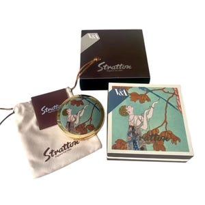 May include: A Stratton compact mirror with a gold-tone frame, featuring an art nouveau design of a woman reaching for leaves. Includes a matching box, a small pouch, and a tag, all bearing the Stratton logo.