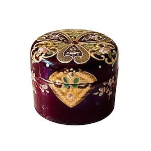 Puede incluir: Una caja decorativa redonda de color burdeos con tapa. La caja está adornada con diseños florales dorados y verdes, y pequeñas flores blancas. La tapa presenta un intrincado patrón dorado con un acento rojo central. La caja probablemente se utiliza para guardar joyas o objetos pequeños.