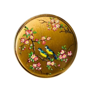 May include: A circular gold-toned compact case featuring a painted design of two small blue and yellow birds perched on brown branches with pink cherry blossoms and green leaves. The metallic surface has a smooth, reflective finish.