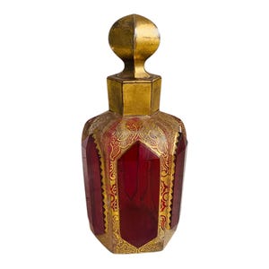 Puede incluir: Un frasco de perfume decorativo con un cuerpo facetado de color rojo rubí y un tapón dorado. La botella presenta diseños de filigrana dorada ornamentada alrededor de los paneles rojos y en el tapón. La botella tiene una forma geométrica y una estética vintage.