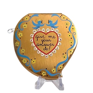 May include: A heart-shaped gold compact mirror featuring a central red heart outline with the text "give me your answer do!" surrounded by a white scalloped border, two blue birds, and yellow floral accents on a metallic gold background.