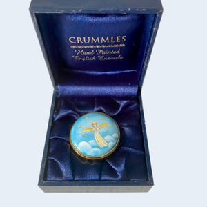 May include: A round Crummles hand painted English enamel box featuring an angel with golden wings flying among blue clouds and yellow stars. The box is presented in a dark blue hinged case with satin lining and the text Crummles Hand Painted English Enamels.