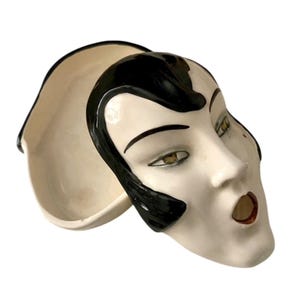 Può includere: Una ciotola in ceramica a forma di volto con base color crema e dettagli neri. Il volto ha un design stilizzato con capelli neri, sopracciglia scure e labbra rosse. La ciotola misura circa 20 cm di lunghezza.