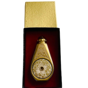 May include: A gold-toned metal perfume bottle with a tapered shape and a circular center inlay featuring an intricate floral engraving. The bottle is housed in a black rectangular box with a red velvet interior lining and a gold-colored lid.