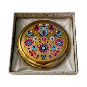 May include: A round gold-toned metal compact mirror featuring a vibrant, multi-coloured floral pattern on the lid. The compact is presented inside a square box with a white fabric lining and a decorative cream-coloured exterior pattern.
