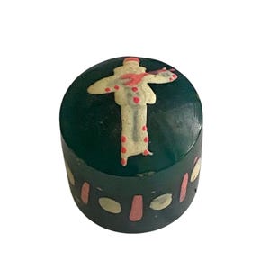 May include: A cylindrical dark green container with a flat top. The lid features a cream-colored figure with red accents. The side of the container is decorated with a repeating pattern of cream-colored ovals and vertical pink rectangular shapes.