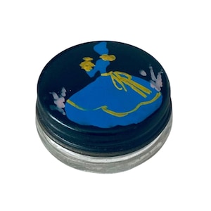 May include: A small, round, clear glass jar with a dark blue lid. The lid features a hand-painted design of a figure in a blue dress with yellow accents, surrounded by small, light pink floral-like brush strokes on the dark blue background.