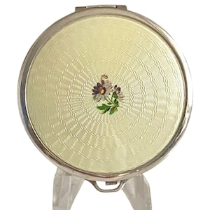 May include: A circular, silver-coloured compact mirror with a pale yellow, textured surface. A small floral design is in the centre. The compact is displayed on a clear, plastic stand.