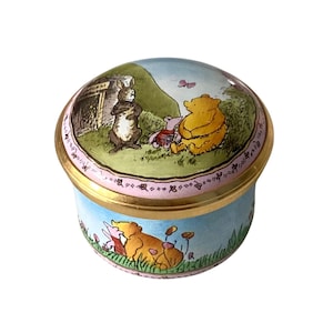 Pode incluir: Uma caixa de joias redonda de porcelana com borda de metal dourado. A tampa apresenta uma ilustração colorida do Ursinho Pooh, Leitão e Coelho em uma paisagem gramada. As laterais da caixa mostram Pooh e Leitão sentados em um campo de flores.